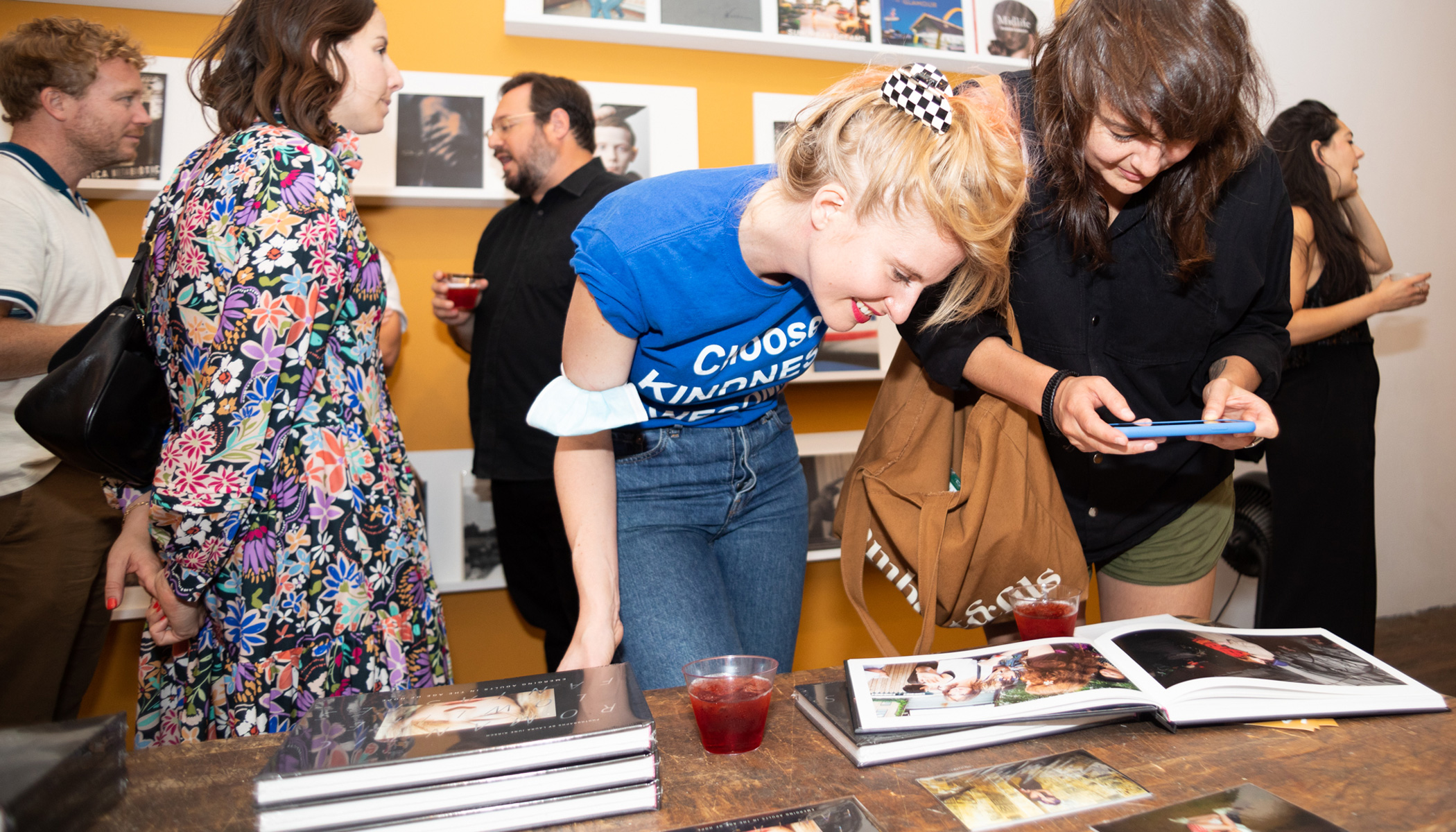 Woman in a blue shirt bent over a table looking at pictures in a book that's on display. Another woman next to her takes a picture of the book on her phone. People in the background walk past and look at the other books on display.