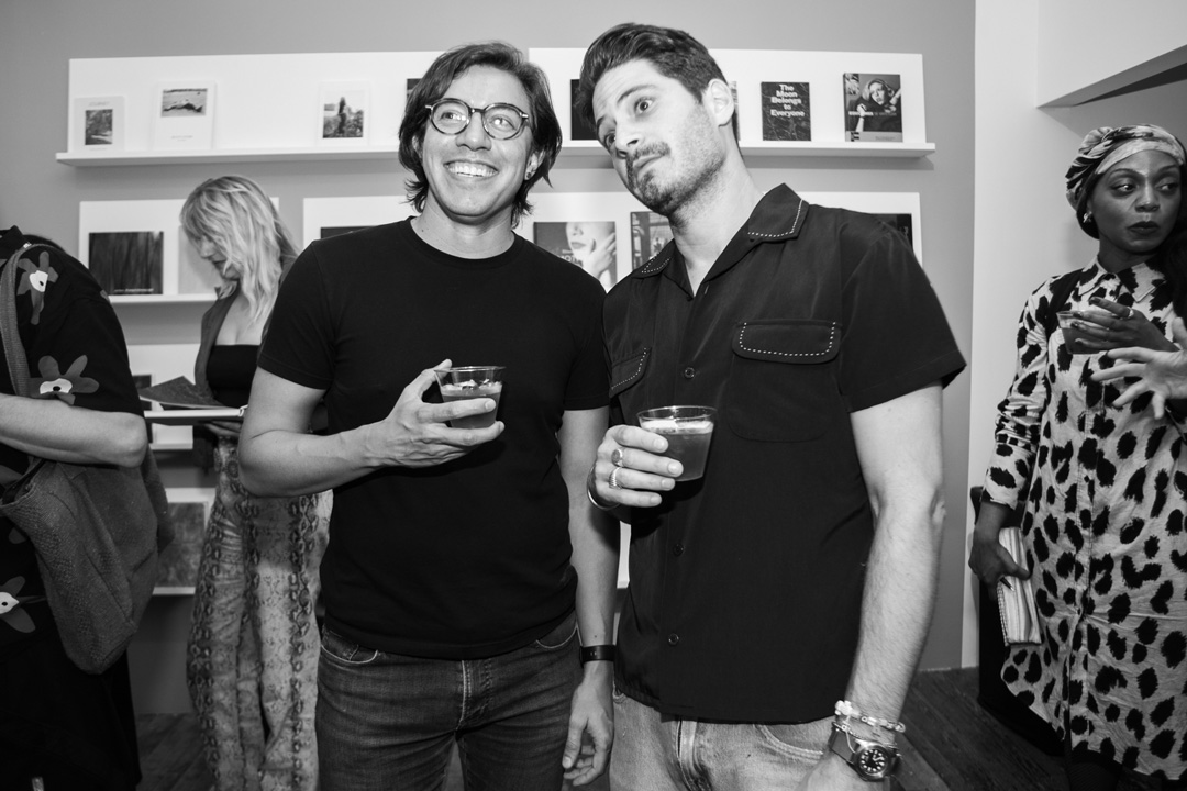 Two men smiling for a photo together with drinks in their hands. They stand in front of a wall with photos on display and a group of people walk around behind them.