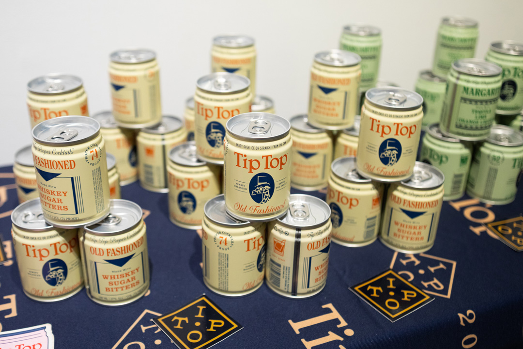 Table display of canned cocktails from Tip Top. These yellow cans are arranged in stacks on a blue table with some green cans next to them on the right and stickers on the table.