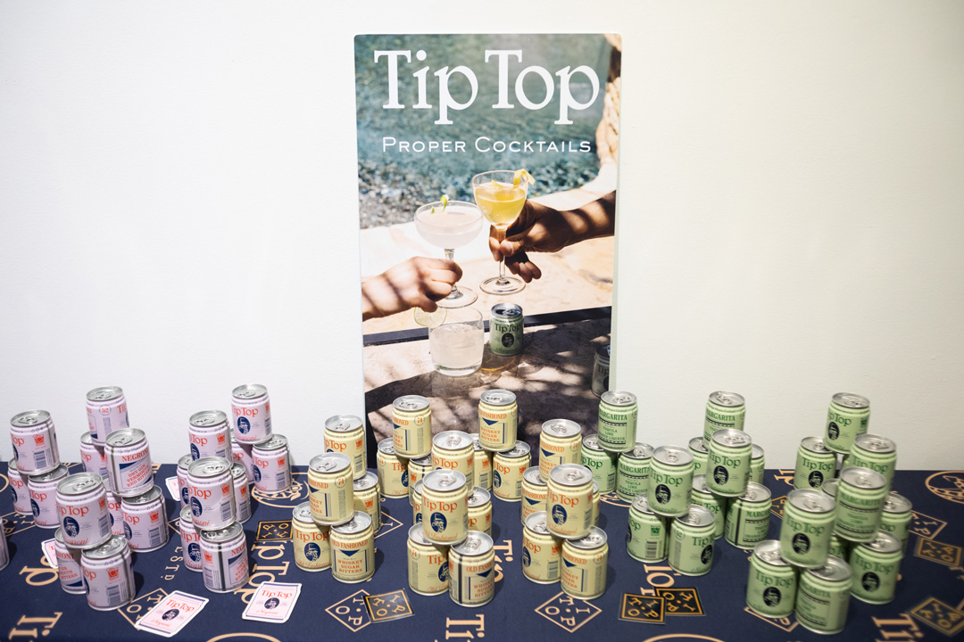 Table display of canned cocktails from Tip Top. Pink cans are stacked on the left, yellow in the middle, and green on the right. There is an advertising poster for the drinks on the wall behind them.