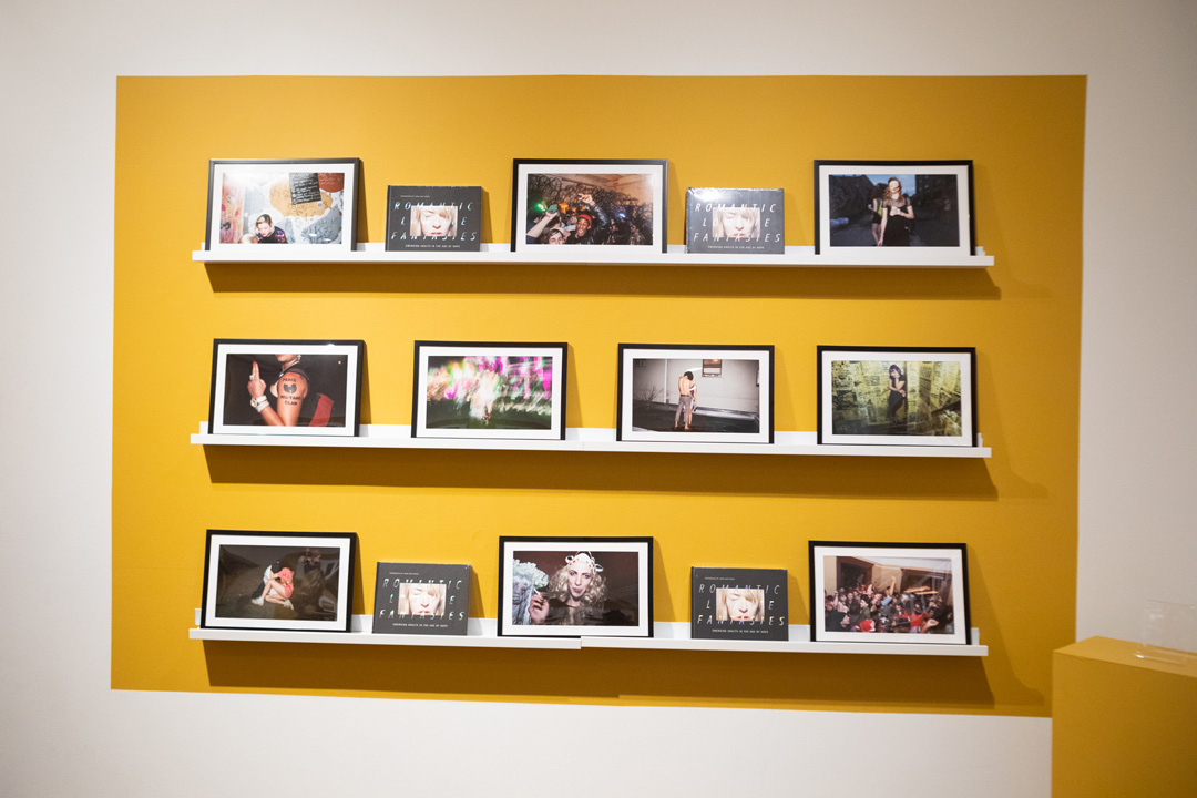 A collection of photos on display on a white and yellow wall. Fourteen photos are on display in small picture frames along the wall.