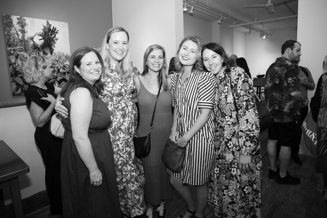 Group of five women smiling for a picture together. They are in an art gallery with other people walking around in the background.