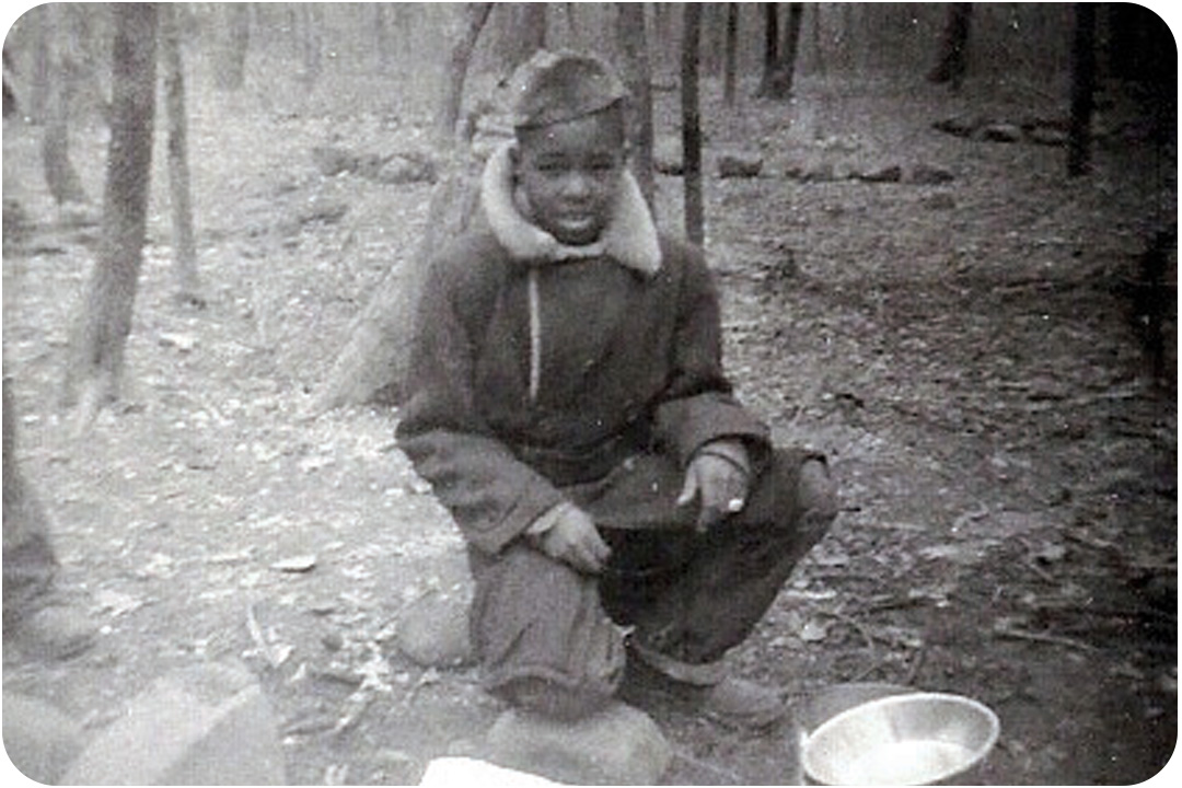 J.R. Harris as a young child camping in the woods as a boy scout. He's wearing a winter coat and a boy scout's cap in the middle of the woods in front of a campsite.