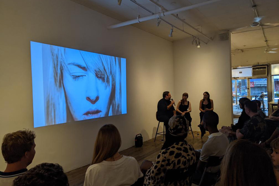 A group of people are sitting in an art gallery while three people sit at the front of the room on high chairs with a microphone. A projector displays a large photo of a girl with blond hair on the wall in front of the group.