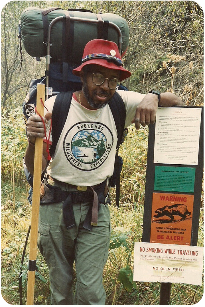 J.R. Harris as a young adult backpacking through Glacier National Park. He is wearing a wilderness explorers t-shirt and his iconic red hat. He's also carrying a large backpack filled with camping gear and holds a walking stick in his hands. He poses next to a trail sign with warning flyers about smoking, fires, and bears.