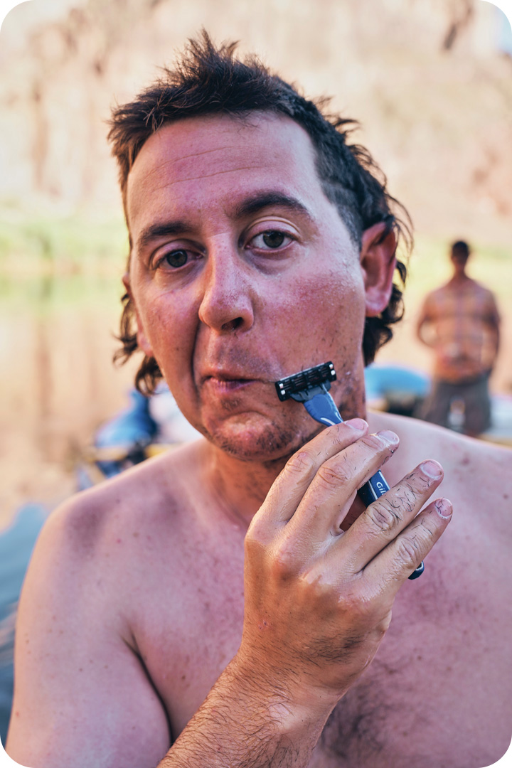 A man shaving his beard with a small razor on the bank of the Colorado River. He looks into the camera and in the background there is another man watiing by the rafts.