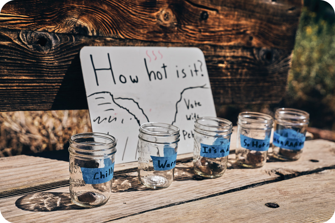Five mason jars sit on a wooden bench all labeled with blue tape with words describing the temperature outside. A dry erase board is propped up behind them all with the words "How hot is it?" written on it.