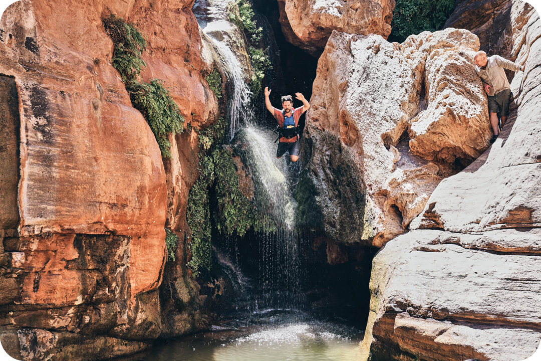 A man jumping off of a tall rock formation into the water below. Behind him is a flowing waterfall and a wall of orange rocks. An older man peers at hime from behind a boulder up above.