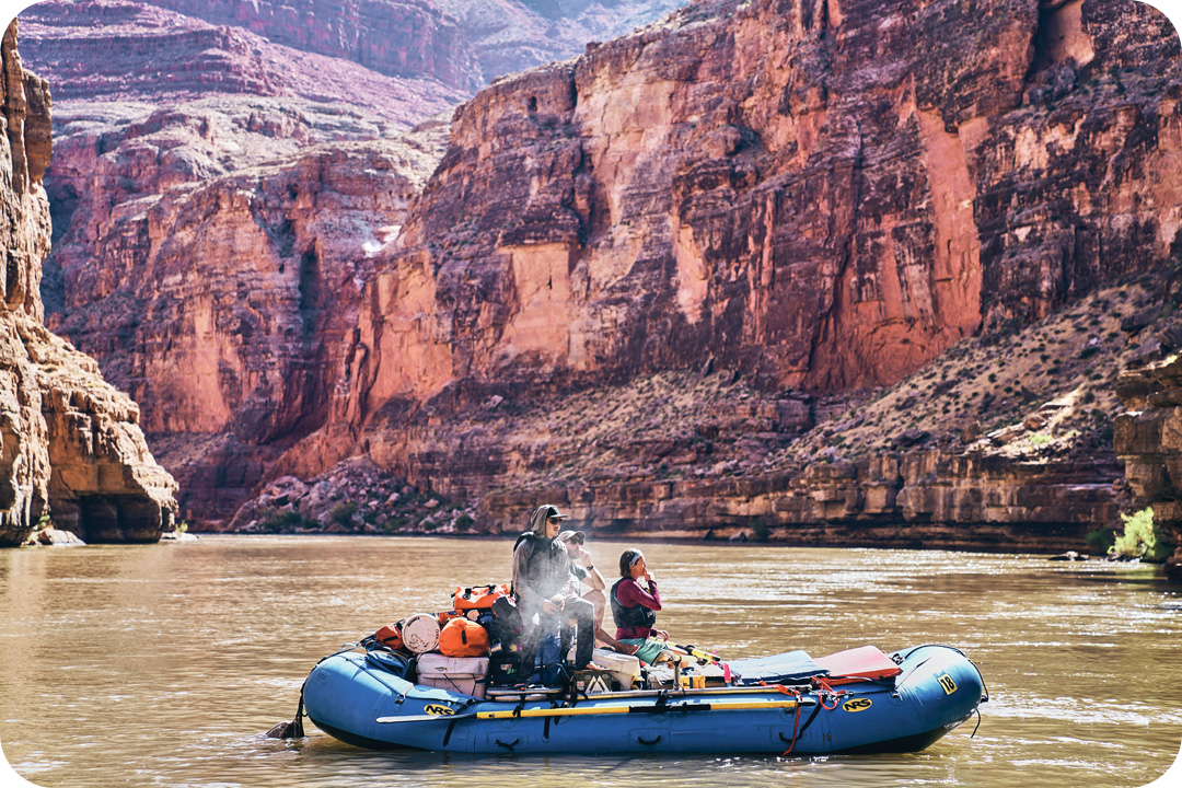 A raft floating down the Colorado River that is filled with supplies and three people. The Grand Canyon is in the background of the image.