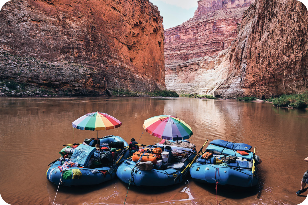 Three river rafts tied to a bank on the Colorado River in the Grand Canyon. All the blue rafts are packed full of supplies, oars, and life preservers. Two of the rafts have an open rainbow umbrella strapped to them.