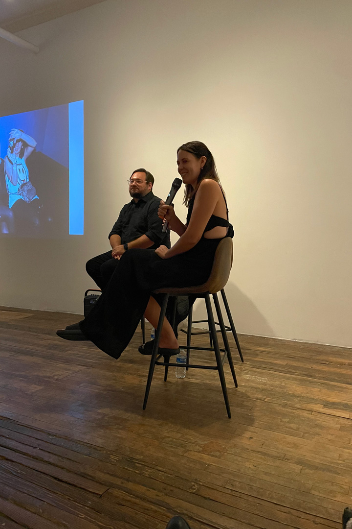 A woman and man sit on high chais at the front of a room while a projector shows photos on the wall behind them. The woman holds a microphone to her mouth and smiles.