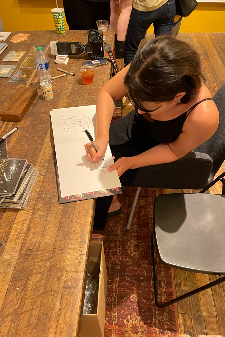 A woman at a table bent over autographing a book. The table is covered in books, pens, and drinks.