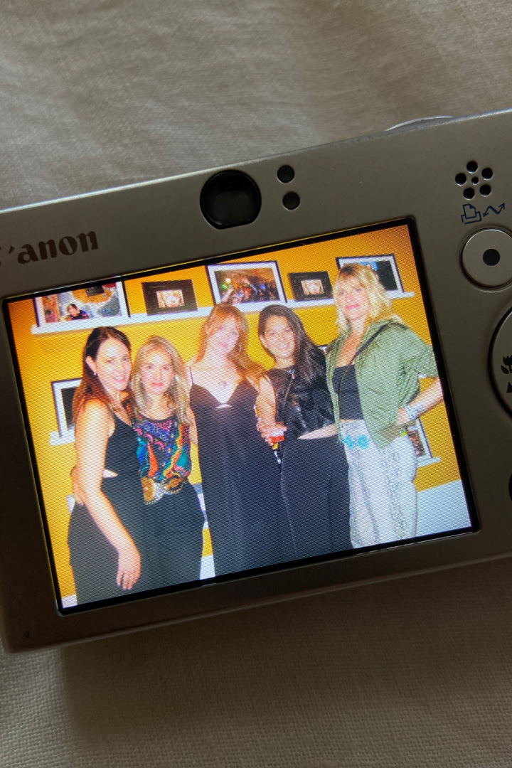 A picture of a digital camera screen that shows a photo of five women smiling together in front of a yellow wall with photos on it.