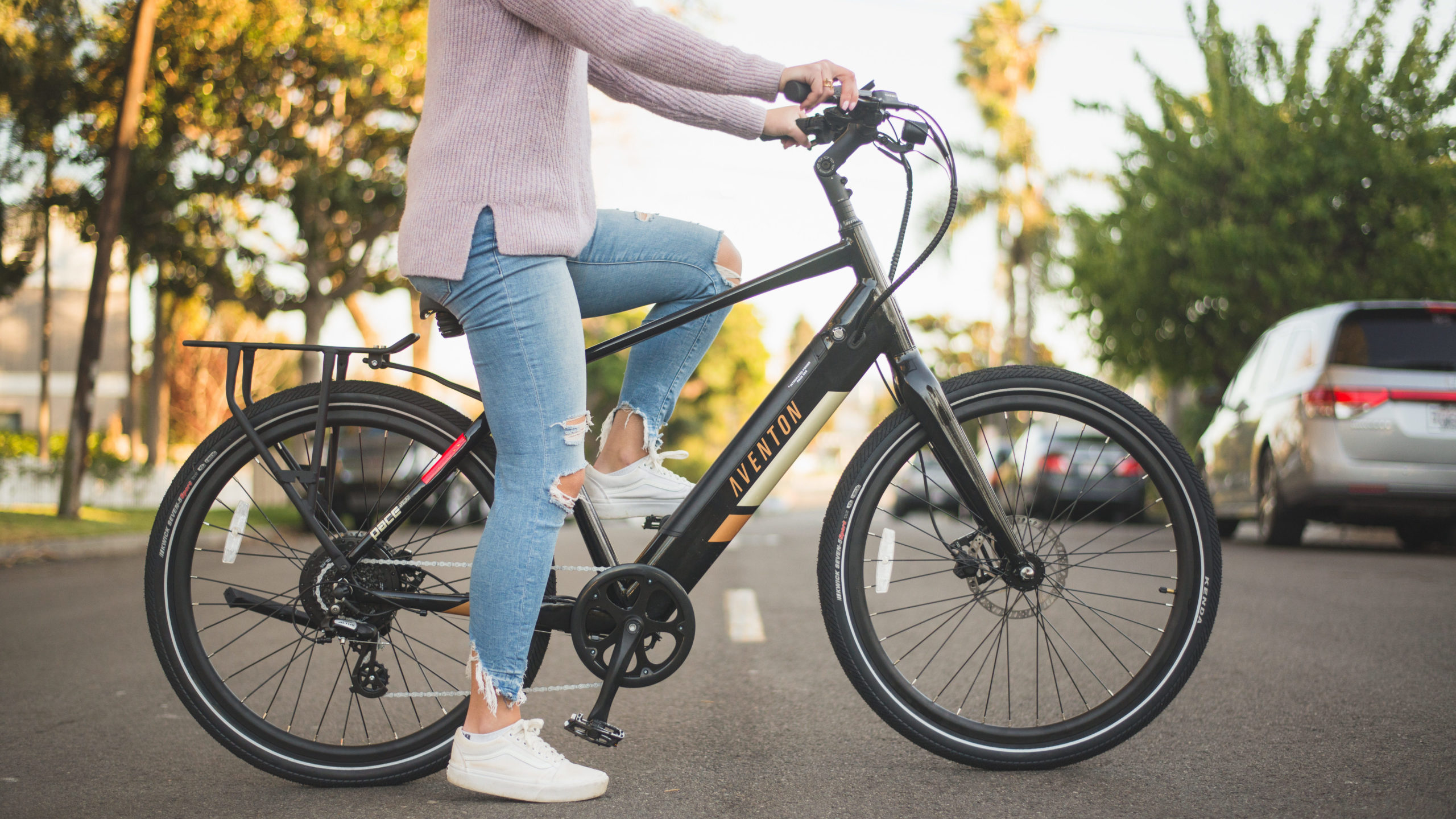 A peson wearing jeans and white sneakers sits on a Midnight Black Pace 500 ebike Next-Gen on the road. Houses and trees and car are in the background.