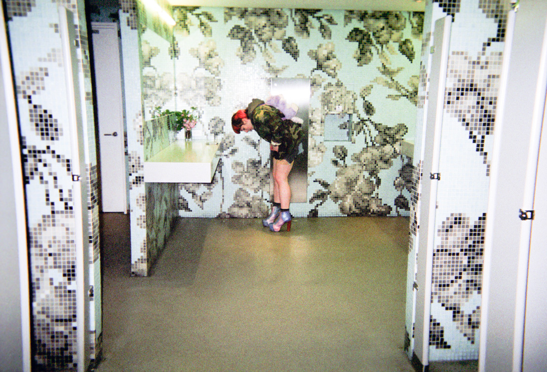 Woman in a bathroom that is covered in black and white floral wallpaper. She is bent over in front of the mirrors and sink adjusting her outfit.