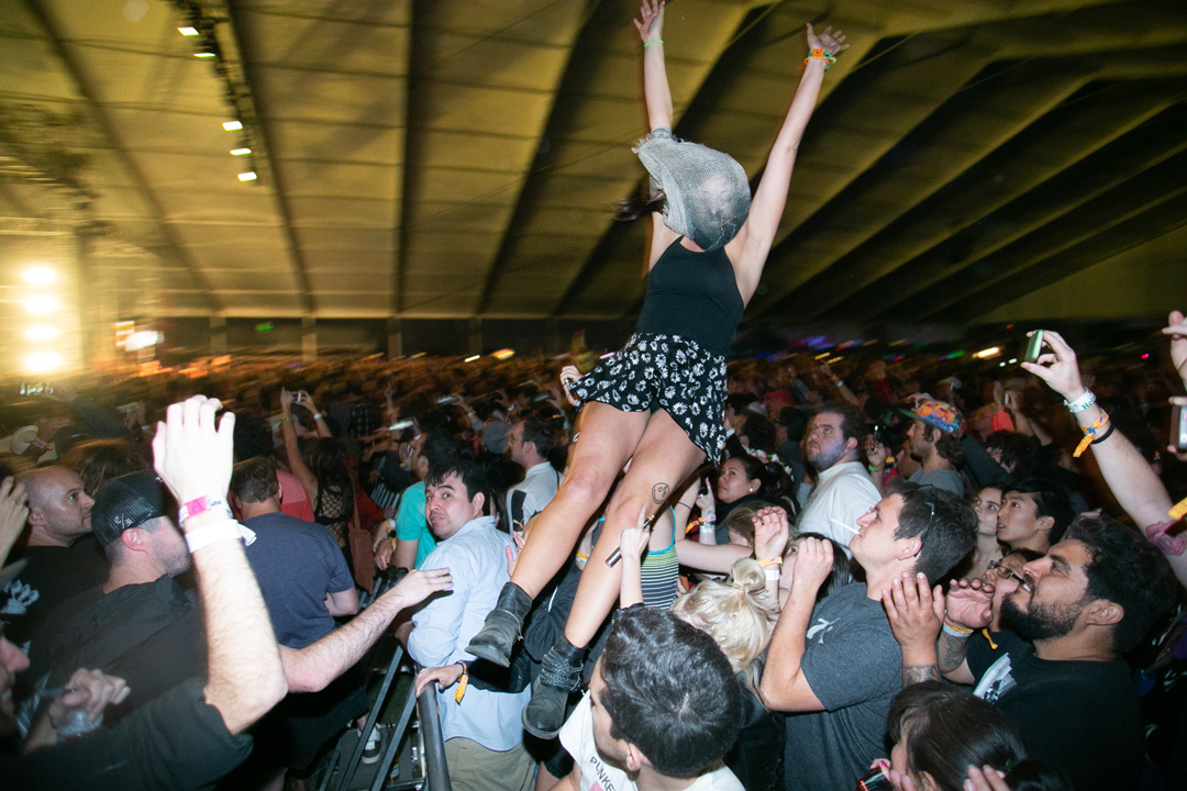 Large group of people at an indoor concert dancing to the music. A woman in a black skirt and top has a mesh bag over her head and is being tossed through the air into the crowd.