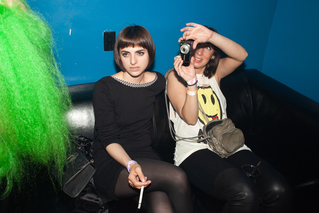 Two girls sitting on a leather couch together looking at something left of the camera. The girl on the left is wearing a black dress and holds a cigarette in her hand while the woman on the right is wearing a white shirt with a yellow smiley face on it and holds up a digital camera to take a picture.