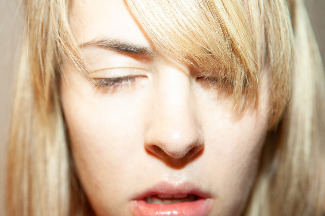 Close up portrait of a woman with blonde hair. Her eyes are closed, mouth slightly open, and her bangs falls sideways over her eyes.