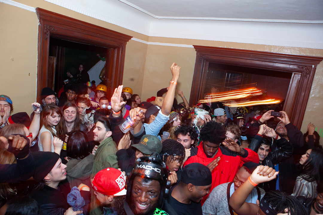 Large group of people at a house party squeezed inside of a room. People are dancing and holding bottles of alcohol and drinks. You can see people coming down a staircase and on other rooms through the open doorways.
