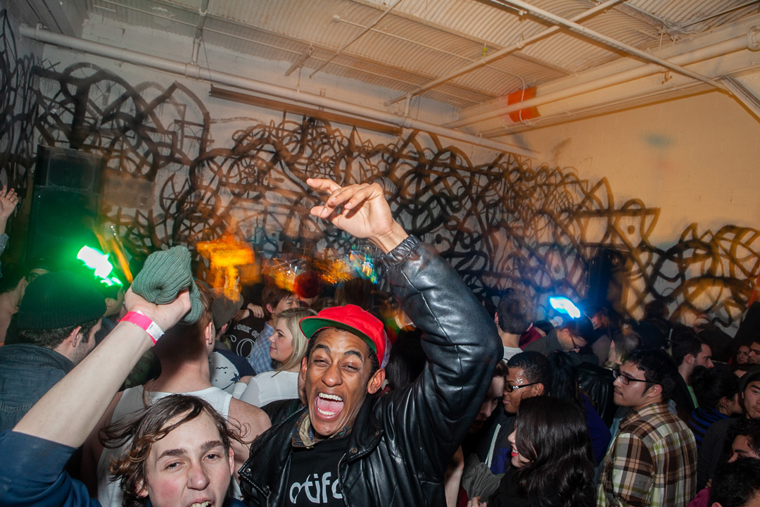 Large group of people in a room with graffiti covering the walls. The center of the image is a man in a leather jacket and red had with his arm above his head and his mouth is open.