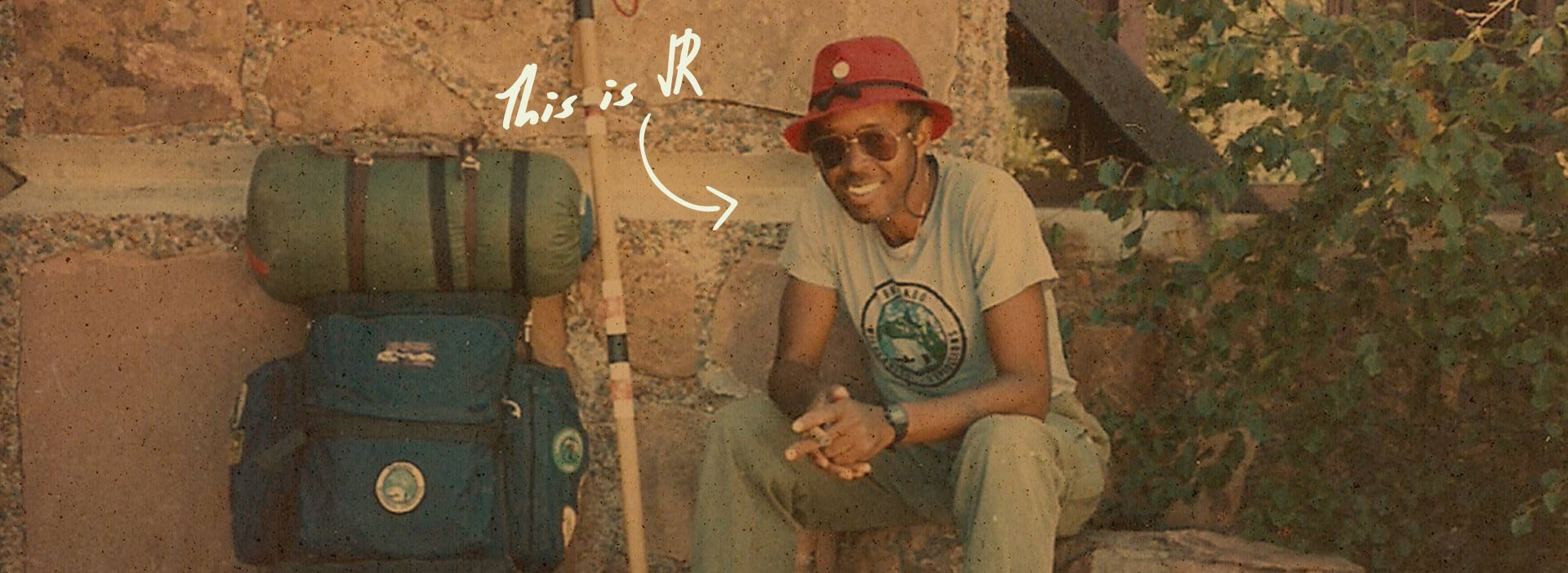 JR Harris sits on a stone ledge next to a stone wall. He's leaning forward and smiling, elbows resting on his knees. He wears a red hat, sunglasses, tshirt, and pants. Next to him, resting against a stone wall is a backback and walking stick.