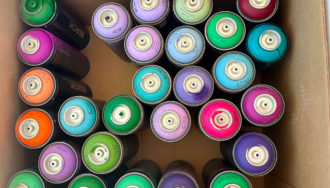 Cardboard box filled with colorful cans of spray paint in shdes of pink, purple, blue, green, and orange.