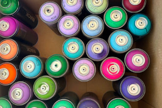 Cardboard box filled with colorful cans of spray paint in shdes of pink, purple, blue, green, and orange.