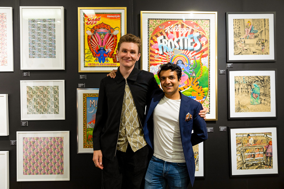 Two people smiling for a picture in front of a gallery wall covered in images. The person on the left wears a black jacket over a green patterned shirt and black pants and the man on the right wear a blue blazer over a white t-shirt and jeans. Behind them there are colorful framed patterns, cartoonish illustrations, and sketchy drawings.