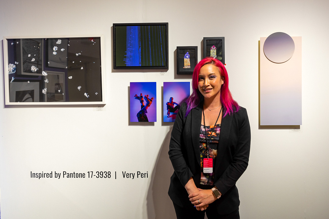 A woman with hot pink hair smiles for a picture in front of a gallery wall of photographs. She wears a black blazer and a colorful, printed shirt underneath. The wall behind her has 7 pieces of art; an abstracts black and white photo, two pictures of someone wearing roller skates, a blue and black piece with blue lines, a rectangle covered in a pink and purple gradient, and two tiny images.