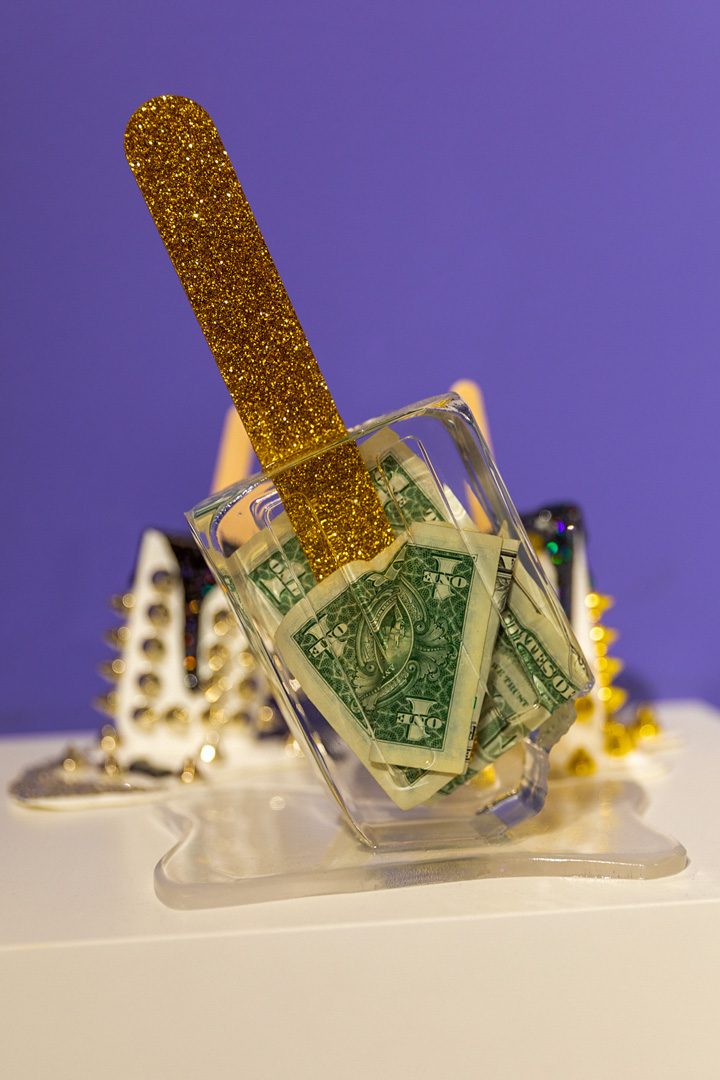 Abstract sculpture sitting on a white shelf. A clear rectangle-shaped container has a gold glitter rounded stick coming out of it and the container is filled with dollar bills. The wall behind the sculpture is a vibrant purple color.