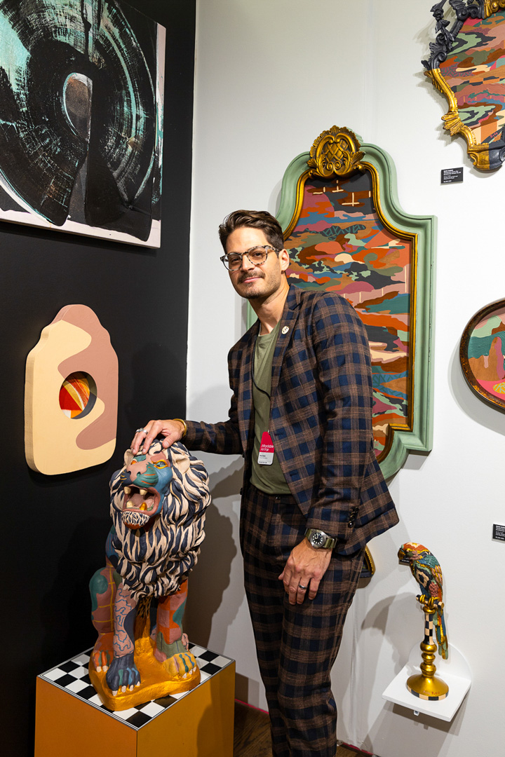 Russ Rubin posing for a picture with a colorfully painted lion sculpture sitting on an orange pedestal with a black and white checkered top. On the walls behind him are colorful paintings in ornate frames.