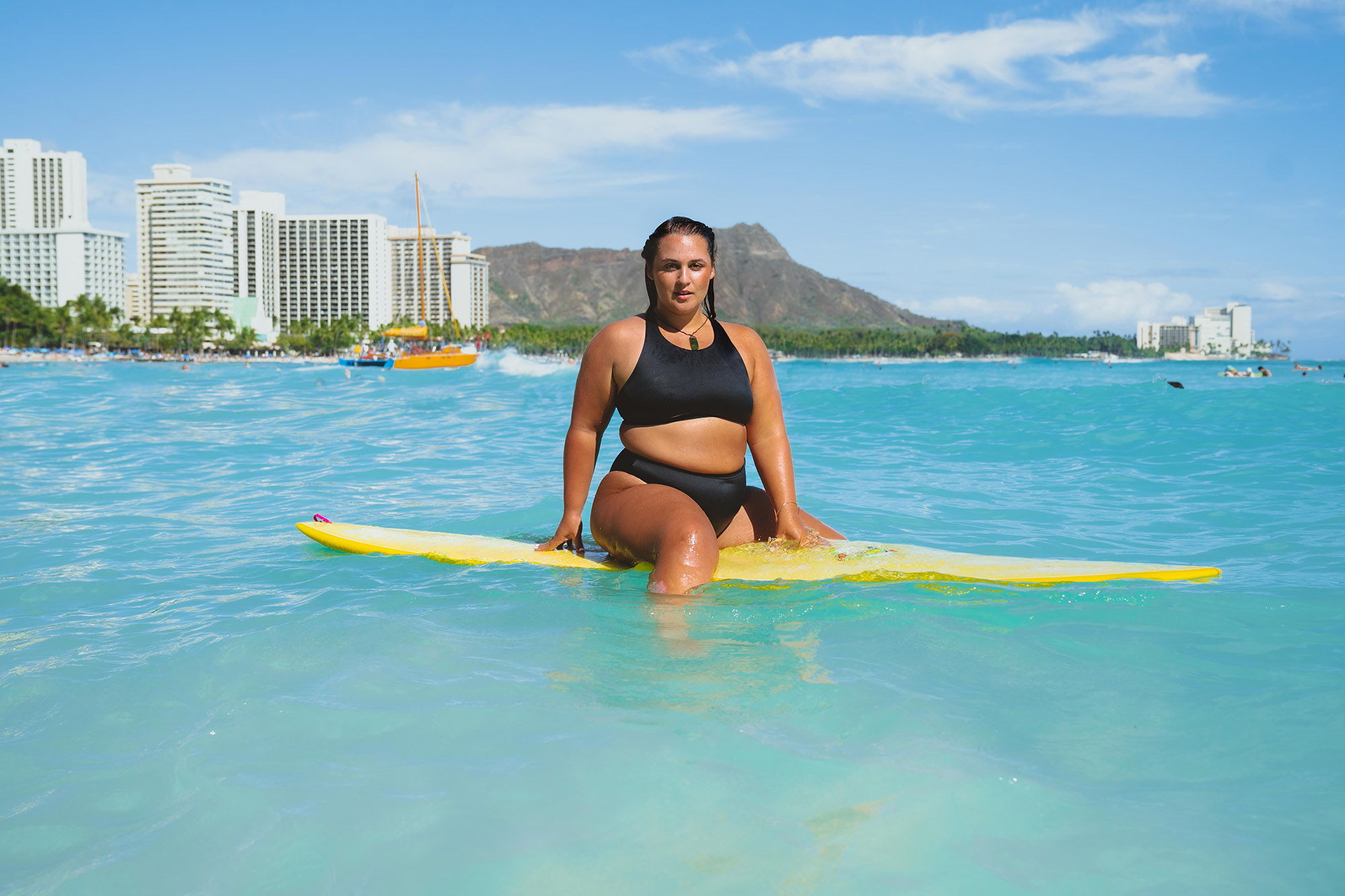 Landscape image of a tan brunette woman in a black two piece swimsuit sitting on a yellow surfboard in an aqua color ocean with a baby blue skyline of white city buildings, brown mountains, and a beach lined with green trees in the background.
