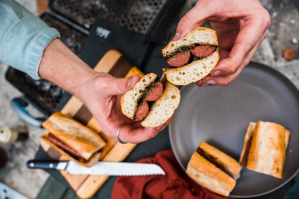 A close up, horizontal feature image of an aerial view of two hands, each holding half of a toasted sub-style sandwich containing two sausage halves cut lengthwise and black garlic chimichurri. In the background, two more sandwich halves are sitting on a dark gray plate in lower right corner. A red cloth napkin is bunched up to the left of the plate. The silver blade of a serrated bread knife is partially laying across the red napkin and the knife’s black handle is resting on a light brown and dark brown striped cutting board in the bottom left corner. A full sandwich is resting on the cutting board above the knife. Part of a black portable charcoal grill is visible in the top left corner.