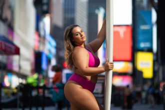 Landscape feature image of a tan, brunette woman in a hot pink two piece bikini posing to the side while holding a white surfboard in front of her on a New York City street filled with people, skyscrapers and brightly lit billboards.
