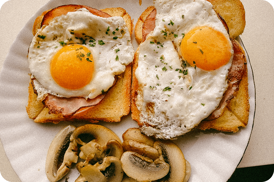 Open faced sandwich on two slices of bread. A piece of meat and over easy egg sit on the bread slices. A piled of cooked mushrooms are next to the sandwich.