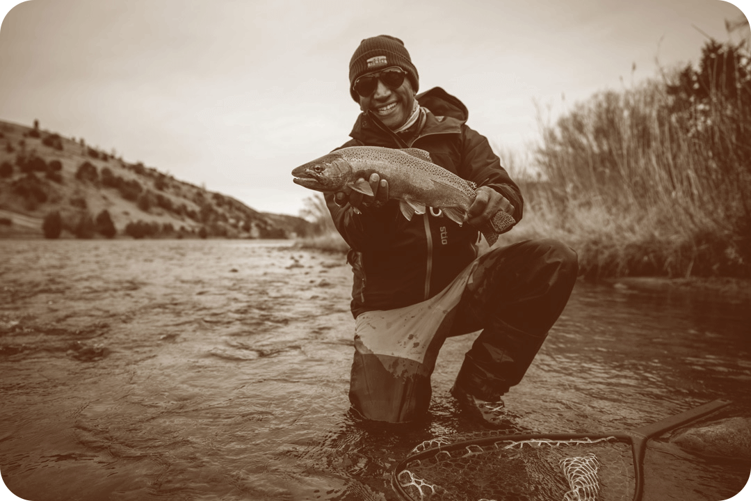 Ranga Perea, chef, posing for a picture holding a fish. Perera is kneeling in the edge of a river in the water and has a large net in front of him. He's wearing waterproof clothing, a beanie, and sunglasses.