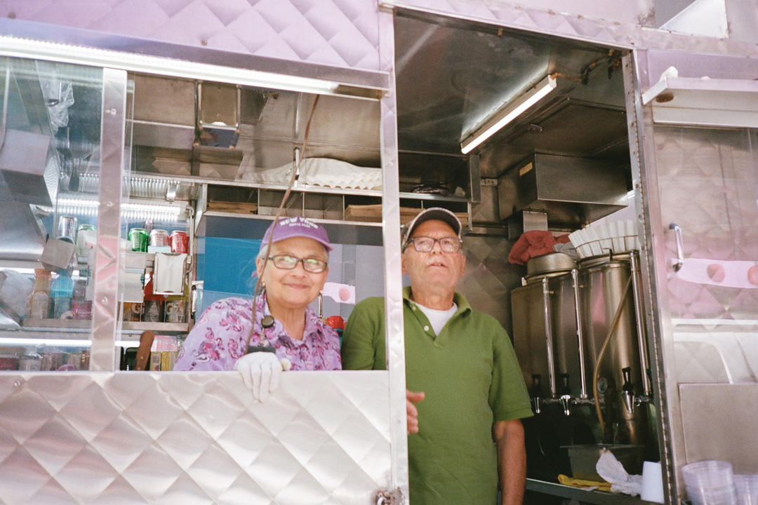 Edith and Angelo Spanoudakis smiling for a picture in the doorway of their breakfast cart. Edith wears a purple patterned shirt and matching hat, Angelo is wearing a green Polo shirt and baseball hat. To the right of the couple are large coffee containers and behind them is a case of cold drinks; Cokes, Sprites, and Gatorades.