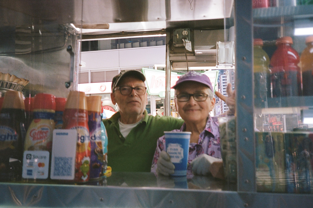 Edith and Angelo Spanoudakis smiling for a picture inside of their breakfast cart. Edith wears a purple patterned shirt and matching hat, Angelo is wearing a green Polo shirt and baseball hat. Edith is handing a cup of coffee out of the window. To the left of the ordering window are stacks of different flavored coffee creamers and to the right is a display case full of Gatorade drinks.