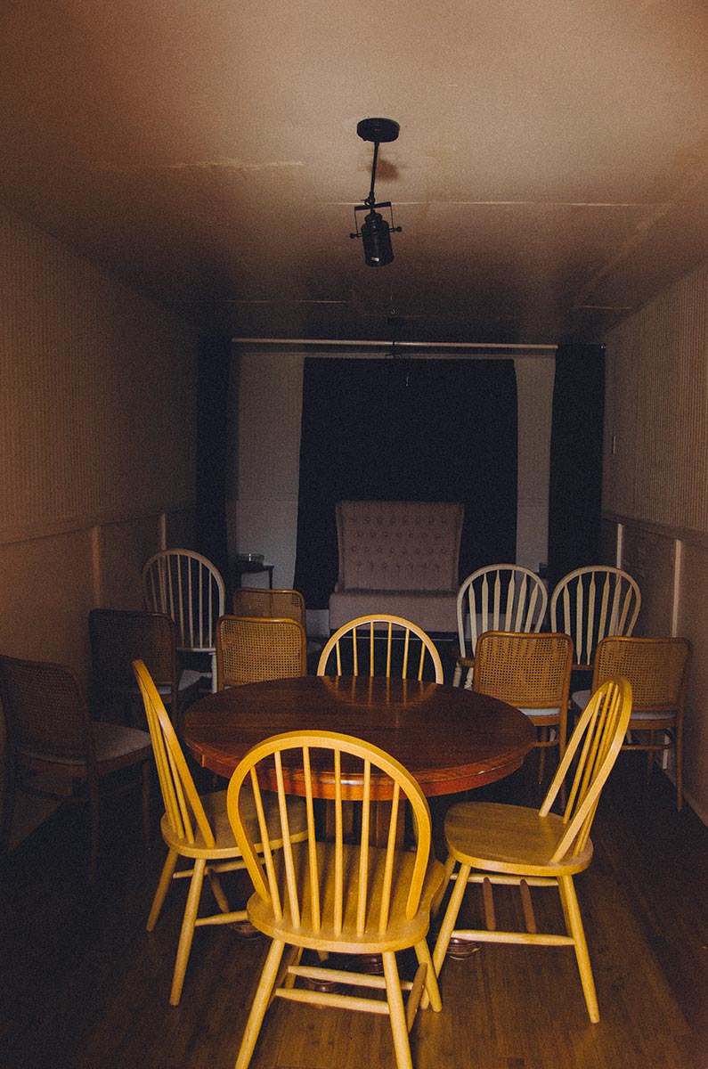 The seance room at the Colby Memorial Temple is dark with a variety of mismatched tables and chairs. An open set of curtains hangs at the back of the room and a large white love seat is against the wall.