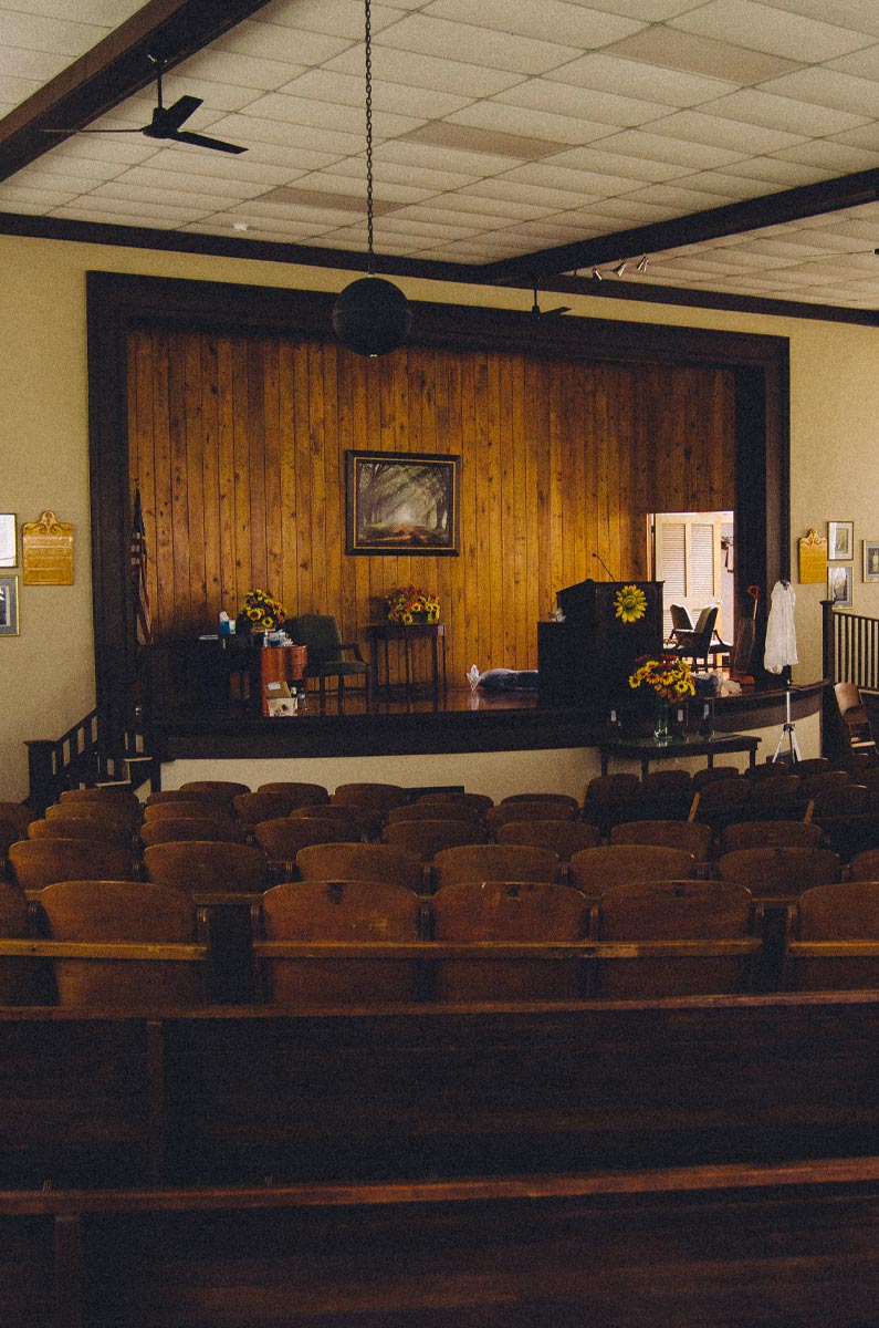 Inside the Colby Memorial Temple are rows of old wood stadium setas and a stage with dark wood paneling behind. Bouquets of flowers are situated around the room. 