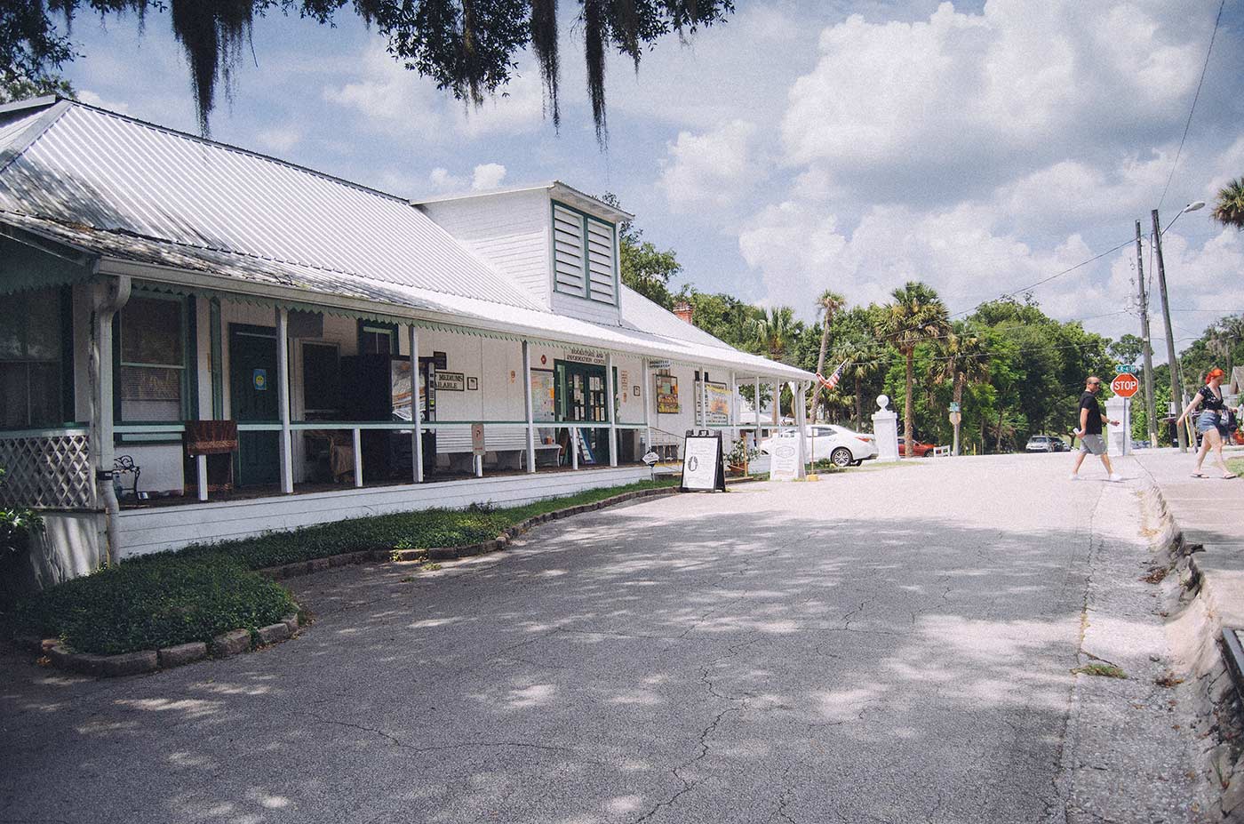 The Cassadaga Welcome Center during the day. Blue sky with clouds overhead. The building has a white metal sloping roof and long porch. it faces a small street. 