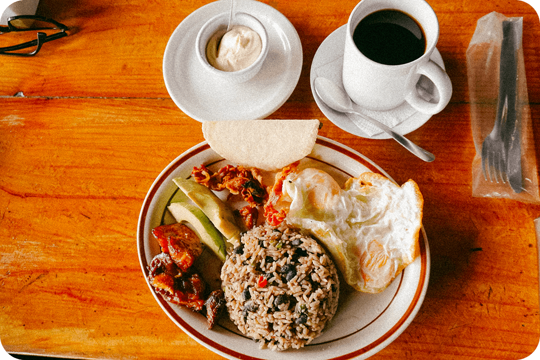 Costa Rican breakfast of gallo pinto (rice and black beans), fried eggs, avocado, and fruit all on a plate. A cup of coffee is sitting next to the play with a spoon laying beside it. A package of silverware is also on the table.