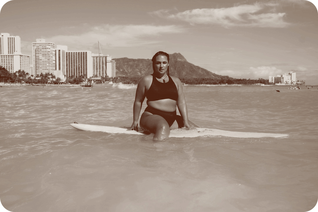 Elizabeth Sneed, surfer, posing for a picture on her surf board in the ocean. She's sitting on her board just offshore and behind her you can see a mountain range and a group of hotel and apartment buildings.