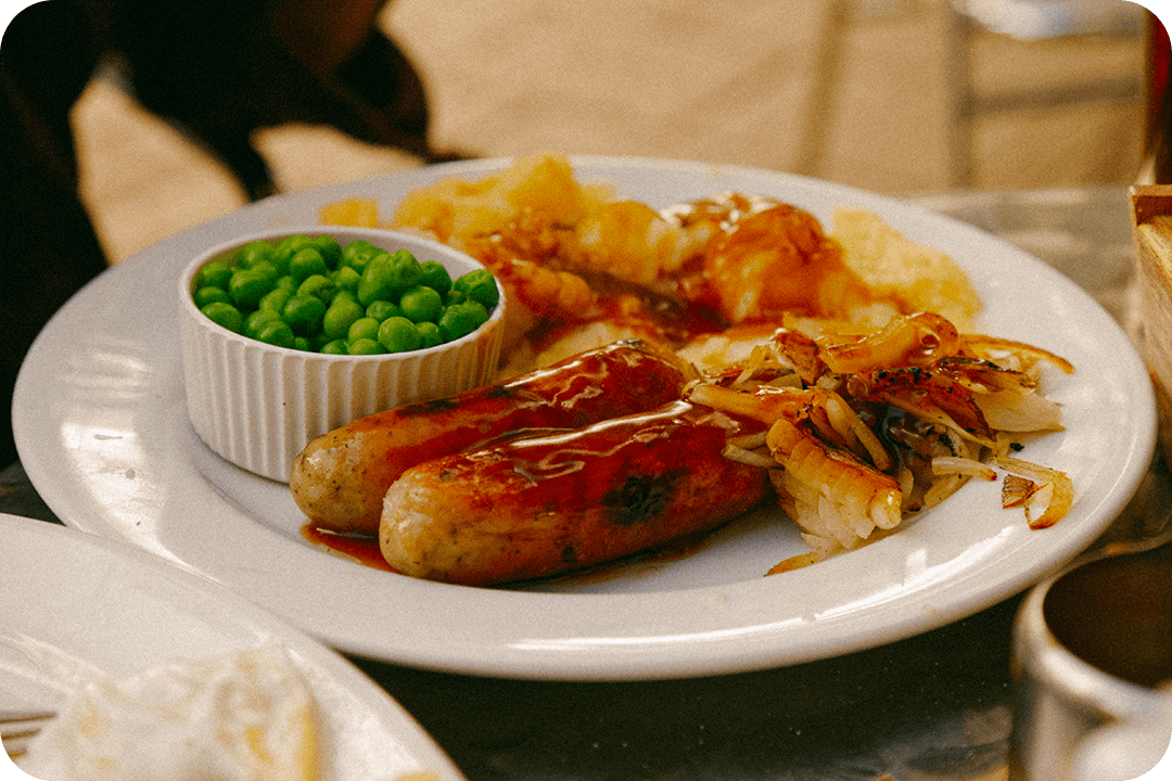 English breakfast of sausages, potatoes, and peas on a white plate sitting on a table.