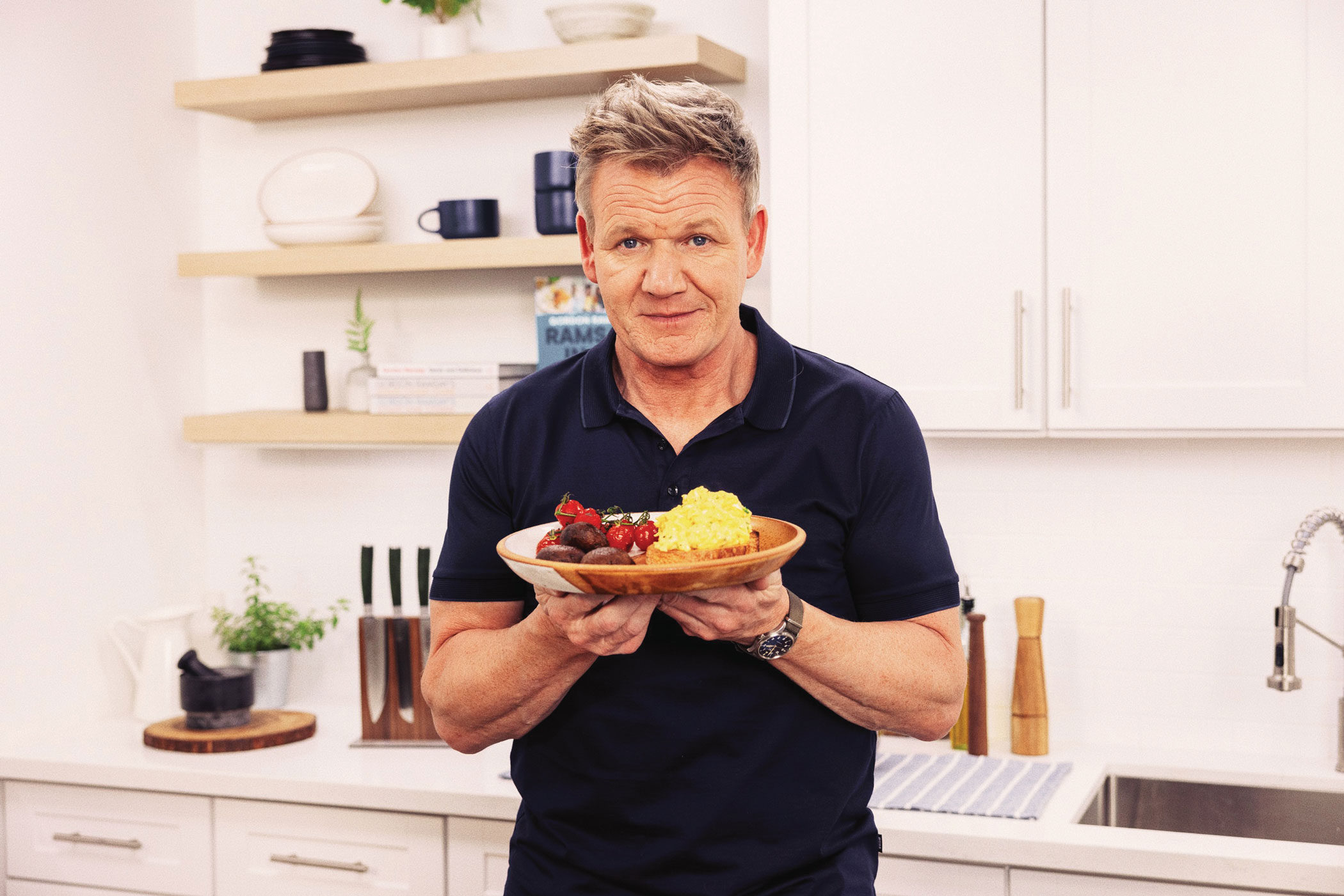Chef Gordon Ramsay in kitchen holding a plate of scrambled eggs.