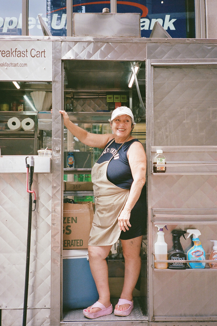 Mama Jo posing for a picture in her breakfast cart. A brown apron covers her black tank top and matching shorts, and she's wearing a white baseball hat and pink sandals. She's standing in the doorway of her cart so you can see into the back.