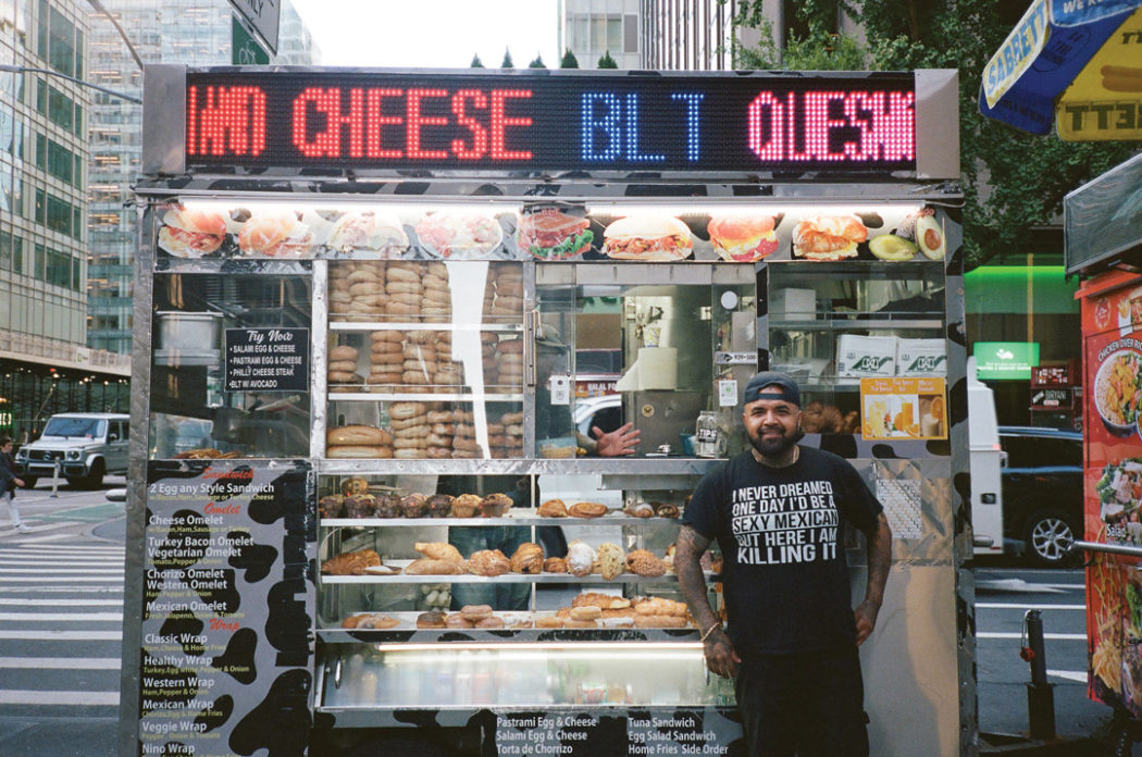 Nino smiling for a picture in front of his breakfast cart on a sidewalk in New York City. The cart has a light-up sign on the top that flashes menu items. Below the sign are pictures of breakfast sandwiches and below that is the ordering window, a glass case full of bagels and pastries, and a menu.