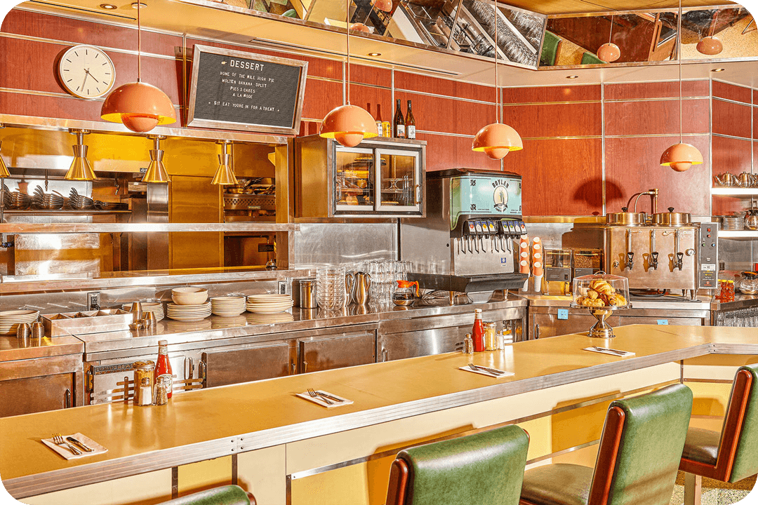 Inside of a diner restaurant. There are four green leather seats pulled up next to a yellow bar top with a set of silverware in front of each seat and a group of condiments. Behind the counter are metal cabinets with stack of white plates, cups, coffee pots, a soda fountain, and a coffee machine. You can see through an opening in the wall back into the kitchen and round lights hang from the ceiling.