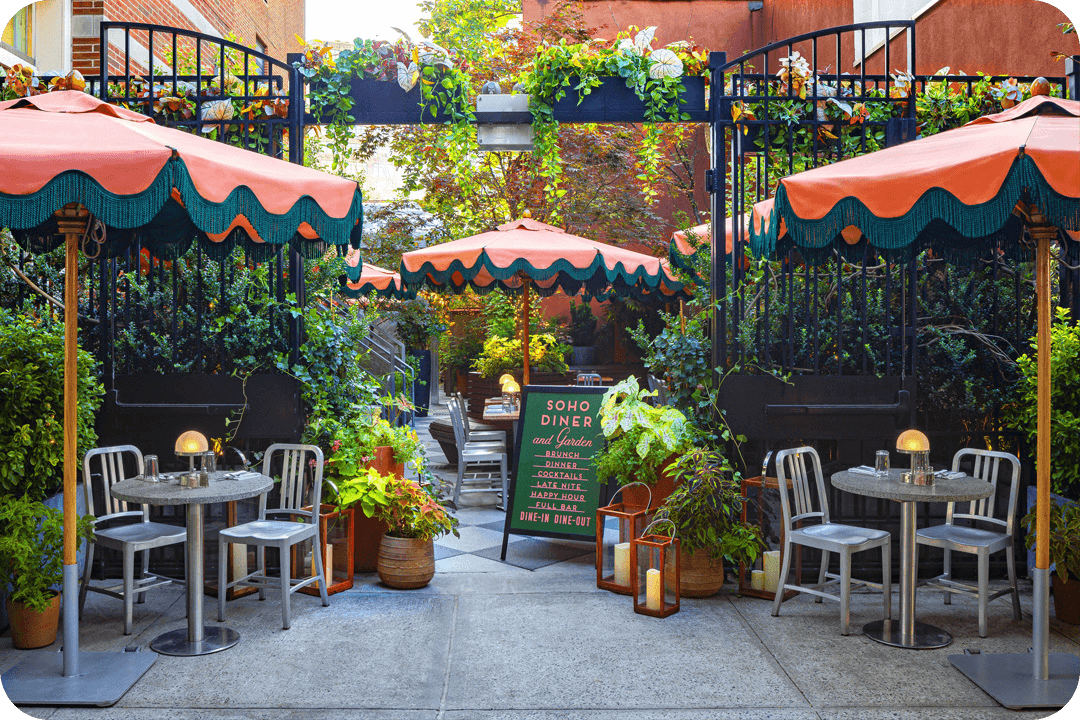 Outside of the Soho Diner. Pink umbrellas with green fringe are set up outside over small metal tables and chairs. A black metal gate opens up into a courtyard filled with more umbrellas and chairs. Greenery covers the metal covers and spills down to the ground and potted plants cover the sidewalk.