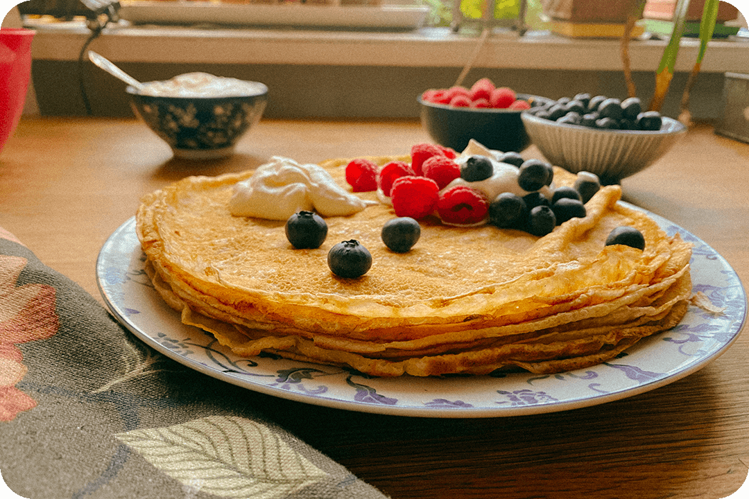 Swedish breakfast of Pannkakor pancakes stacked on a plate. A pile of blueberries, raspberries, and a dollop of cream are sitting on top. On the table behind are two bowls of blueberries and raspberries.
